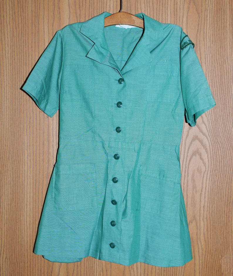 May include: A vintage green Girl Guide uniform dress with short sleeves and a button-up front. The dress has two pockets and a patch on the right sleeve.