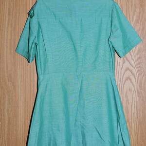 May include: A green short-sleeved shirt with a collared neckline and a button-up front. The shirt has a flared skirt and is made of a lightweight fabric.
