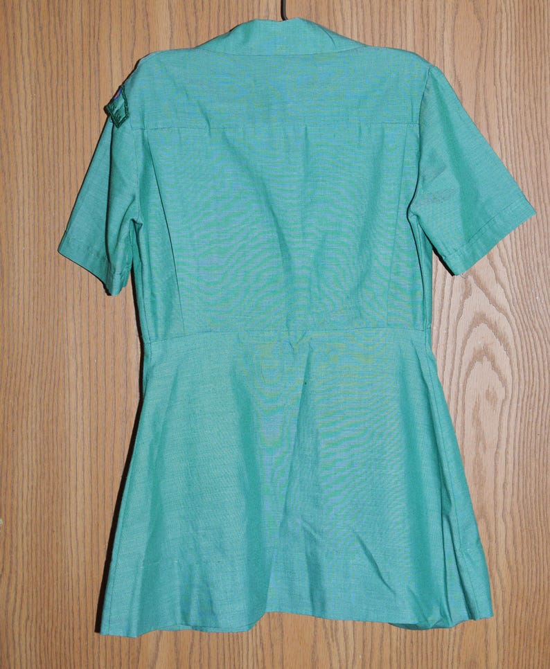 May include: A green short-sleeved shirt with a collared neckline and a button-up front. The shirt has a flared skirt and is made of a lightweight fabric.