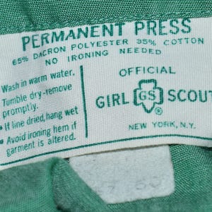 May include: A white tag with green text that reads "PERMANENT PRESS 65% DACRON POLYESTER 35% COTTON NO IRONING NEEDED" and "OFFICIAL GIRL GS SCOUTS NEW YORK, N.Y.". The tag also includes care instructions: "Wash in warm water. Tumble dry-remove promptly. If line dried, hang wet. Avoid ironing hem if garment is altered."