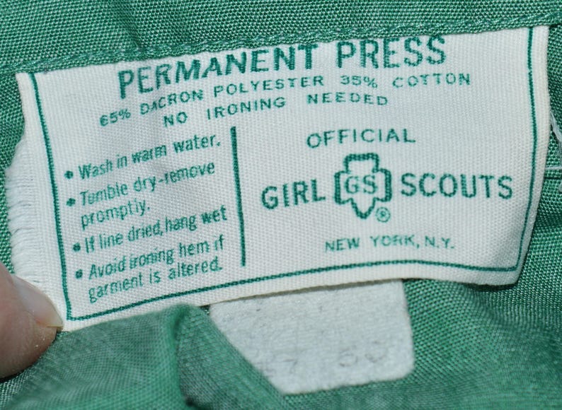 May include: A white tag with green text that reads "PERMANENT PRESS 65% DACRON POLYESTER 35% COTTON NO IRONING NEEDED" and "OFFICIAL GIRL GS SCOUTS NEW YORK, N.Y.". The tag also includes care instructions: "Wash in warm water. Tumble dry-remove promptly. If line dried, hang wet. Avoid ironing hem if garment is altered."