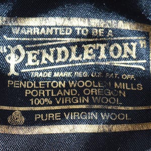 Puede incluir: Primer plano de una etiqueta negra con letras doradas. La etiqueta dice "PENDLETON" e incluye texto sobre el origen de la marca en Portland, Oreg&oacute;n, y el uso de 100% lana virgen. La etiqueta es rectangular.