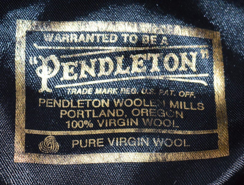 Puede incluir: Primer plano de una etiqueta negra con letras doradas. La etiqueta dice "PENDLETON" e incluye texto sobre el origen de la marca en Portland, Oreg&oacute;n, y el uso de 100% lana virgen. La etiqueta es rectangular.