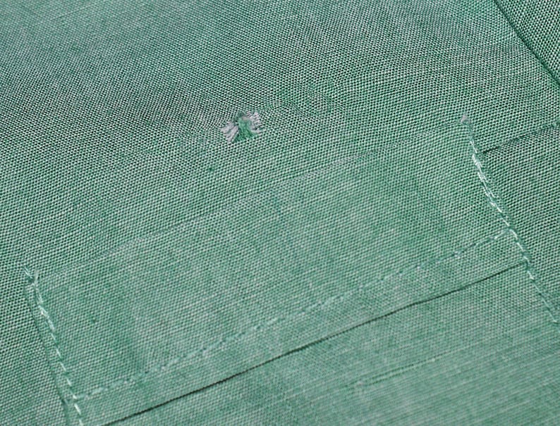 May include: A close-up of a green fabric with a small hole in the centre. The fabric has a pocket with a stitched edge.