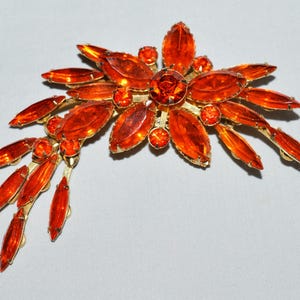 May include: A vintage brooch featuring a floral design with orange-colored, oval-shaped rhinestones and smaller round stones. The brooch has a gold-tone metal setting. The brooch is approximately 3 inches wide.
