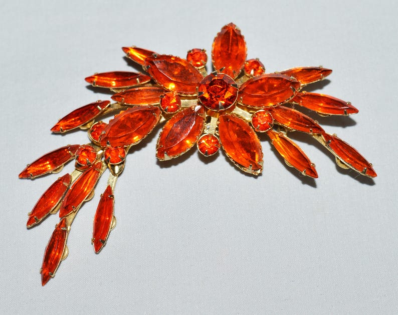 May include: A vintage brooch featuring a floral design with orange-colored, oval-shaped rhinestones and smaller round stones. The brooch has a gold-tone metal setting. The brooch is approximately 3 inches wide.