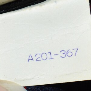 Puede incluir: Primer plano de una etiqueta blanca con el texto impreso "A201-367" en tinta azul. La etiqueta est&aacute; ligeramente arrugada y parece estar adherida a una prenda con tela de cuadros rojos y negros visible.