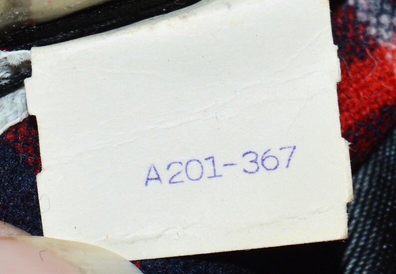 Puede incluir: Primer plano de una etiqueta blanca con el texto impreso "A201-367" en tinta azul. La etiqueta est&aacute; ligeramente arrugada y parece estar adherida a una prenda con tela de cuadros rojos y negros visible.