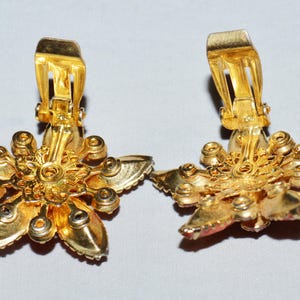 May include: A pair of gold-toned clip-on earrings. Each earring is shaped like a five-petal flower with a central design and small, round accents. The clip-on mechanism is visible on the back. The earrings have a vintage aesthetic.