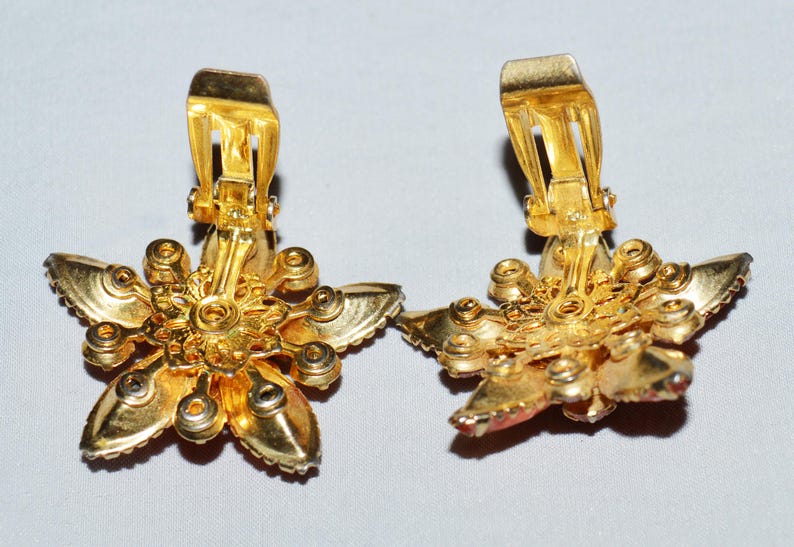 May include: A pair of gold-toned clip-on earrings. Each earring is shaped like a five-petal flower with a central design and small, round accents. The clip-on mechanism is visible on the back. The earrings have a vintage aesthetic.
