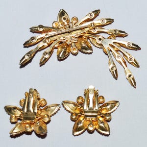 May include: A vintage gold-toned jewelry set featuring a brooch and matching clip-on earrings. The brooch has a starburst design with clear and amber-colored rhinestones. The earrings are flower-shaped with similar rhinestones.