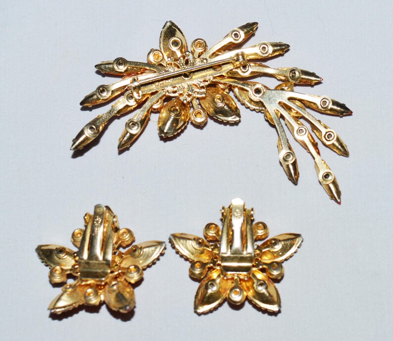 May include: A vintage gold-toned jewelry set featuring a brooch and matching clip-on earrings. The brooch has a starburst design with clear and amber-colored rhinestones. The earrings are flower-shaped with similar rhinestones.