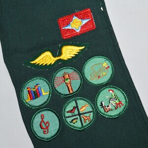May include: Green fabric sash with various embroidered badges, including a red badge with a white star and a blue flower, a yellow wing, a green circle with a red musical note, a green circle with a red and blue book stack, a green circle with a red and white lighthouse, a green circle with a red and yellow bird, a green circle with a red and yellow fish, a green circle with a red and yellow dog, a green circle with a red and yellow person playing a musical instrument, and a green circle with a red and yellow person sitting on a bench.