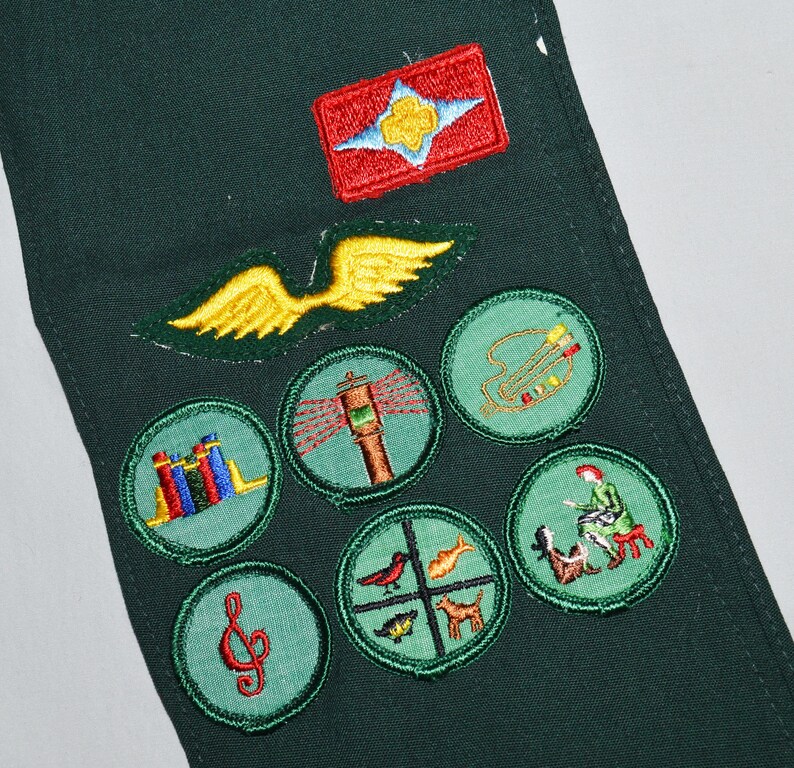 May include: Green fabric sash with various embroidered badges, including a red badge with a white star and a blue flower, a yellow wing, a green circle with a red musical note, a green circle with a red and blue book stack, a green circle with a red and white lighthouse, a green circle with a red and yellow bird, a green circle with a red and yellow fish, a green circle with a red and yellow dog, a green circle with a red and yellow person playing a musical instrument, and a green circle with a red and yellow person sitting on a bench.