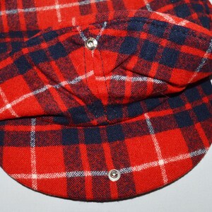 Puede incluir: Gorra a cuadros rojos, azul marino y blancos con visera redondeada. La gorra est&aacute; hecha de tela tejida y tiene dos cierres de bot&oacute;n a presi&oacute;n plateados. La gorra est&aacute; doblada, revelando el forro interior.