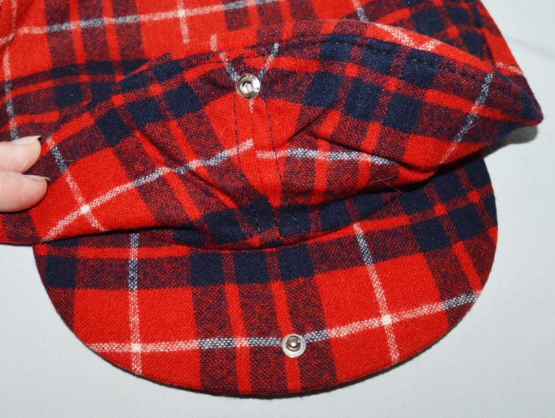 Puede incluir: Gorra a cuadros rojos, azul marino y blancos con visera redondeada. La gorra est&aacute; hecha de tela tejida y tiene dos cierres de bot&oacute;n a presi&oacute;n plateados. La gorra est&aacute; doblada, revelando el forro interior.