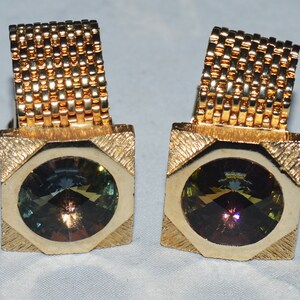 May include: A pair of gold cufflinks with a square design and a round, multi-colored gemstone in the center.