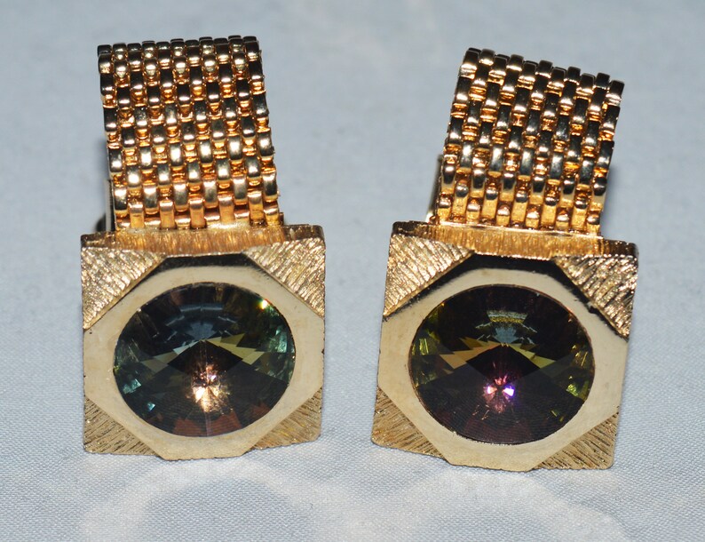 May include: A pair of gold cufflinks with a square design and a round, multi-colored gemstone in the center.