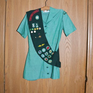 May include: A green Girl Guide uniform with a sash and badges. The sash has the text "Four Winds Council" and the number "81" on it.