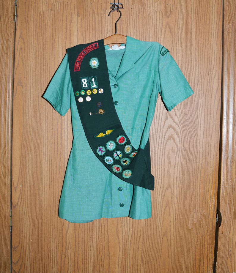May include: A green Girl Guide uniform with a sash and badges. The sash has the text "Four Winds Council" and the number "81" on it.