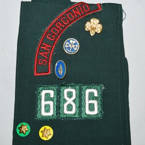 May include: A green Girl Guide sash with a red and white patch that says "SAN GORGOINO" and a number patch that says "686". There are also several small badges, including a gold clover, a blue clover, and a blue hand.