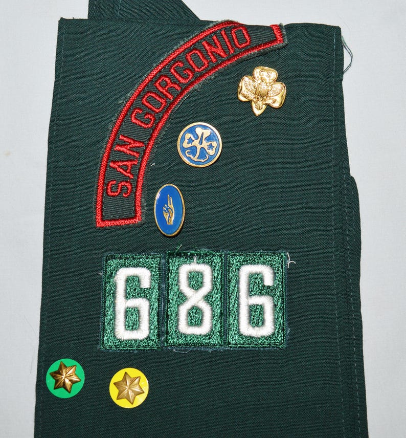 May include: A green Girl Guide sash with a red and white patch that says "SAN GORGOINO" and a number patch that says "686". There are also several small badges, including a gold clover, a blue clover, and a blue hand.