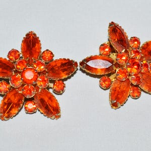 May include: A pair of vintage flower-shaped clip-on earrings. Each earring features a gold-tone metal setting with large, orange-colored, marquise-cut and round-cut rhinestones. The earrings are set against a light gray background.