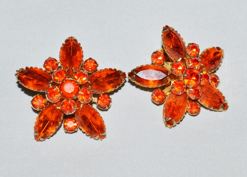 May include: A pair of vintage flower-shaped clip-on earrings. Each earring features a gold-tone metal setting with large, orange-colored, marquise-cut and round-cut rhinestones. The earrings are set against a light gray background.