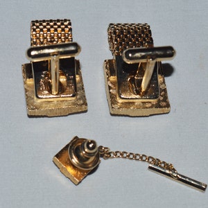 May include: A set of gold-toned cufflinks and a tie tack. The cufflinks have a square design with a textured surface. The tie tack has a similar design and is attached to a chain.