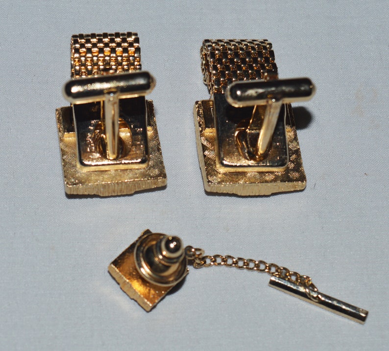 May include: A set of gold-toned cufflinks and a tie tack. The cufflinks have a square design with a textured surface. The tie tack has a similar design and is attached to a chain.