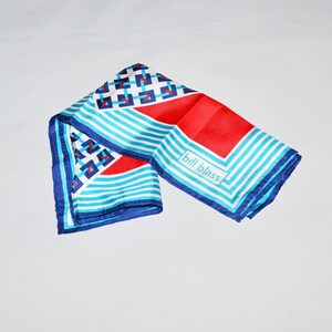 May include: A square scarf featuring a geometric pattern of blue and red squares on a white background, accented by light blue and white stripes. The scarf includes a solid red section and the text bill blass printed in a white rectangular box.