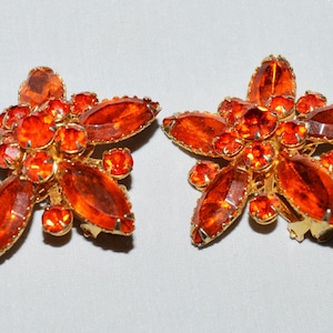 May include: A pair of vintage clip-on earrings. Each earring is shaped like a flower with orange-red rhinestones and a gold-tone metal setting. The earrings have a cluster of smaller stones in the center.
