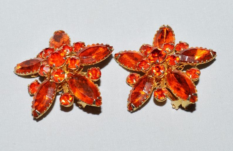 May include: A pair of vintage clip-on earrings. Each earring is shaped like a flower with orange-red rhinestones and a gold-tone metal setting. The earrings have a cluster of smaller stones in the center.