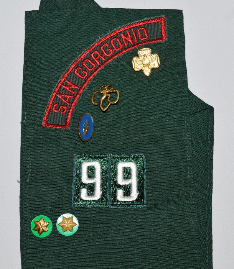 May include: Green Girl Guide sash with a red and green patch that says "SAN GORGONIO", a gold flower badge, a blue badge with a flame design, a gold badge with two overlapping four-leaf clovers, a white number "99" patch, and two green star badges.