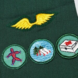 May include: Green fabric with three embroidered badges. The badges are circular and feature a red stick figure, a blue and white scene with a tent, and a white first aid kit with a red cross.