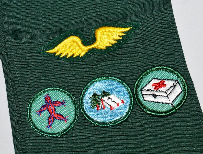 May include: Green fabric with three embroidered badges. The badges are circular and feature a red stick figure, a blue and white scene with a tent, and a white first aid kit with a red cross.