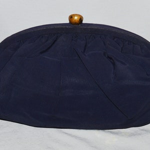 May include: A navy blue fabric clutch purse with a wooden top handle. The purse has a gathered fabric design and a gold chain strap.