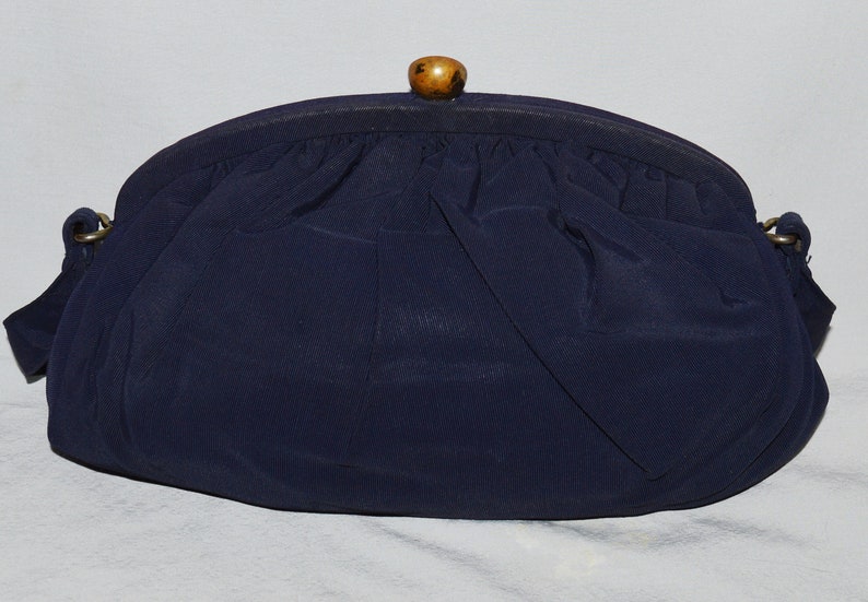 May include: A navy blue fabric clutch purse with a wooden top handle. The purse has a gathered fabric design and a gold chain strap.
