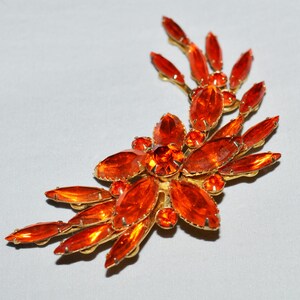 May include: A vintage brooch featuring a floral design with orange-colored, oval-cut rhinestones set in a gold-tone metal. The brooch has a curved, asymmetrical shape, with the rhinestones radiating outwards.