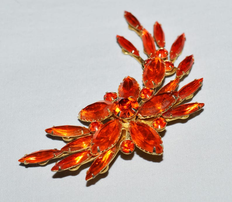 May include: A vintage brooch featuring a floral design with orange-colored, oval-cut rhinestones set in a gold-tone metal. The brooch has a curved, asymmetrical shape, with the rhinestones radiating outwards.