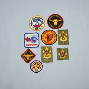 May include: A collection of eight embroidered patches featuring various designs related to scouting. The patches are in different colors, including yellow, red, blue, green, and white. Some of the patches feature text, such as "Follow the Rugged Road", "Quivira Scout Ranch", "Camporee", and "Be Prepared".