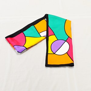 May include: A colorful silk scarf with a geometric pattern. The scarf features a black border and a design of bright colors including pink, yellow, orange, green, and purple. The scarf is folded on a white surface.