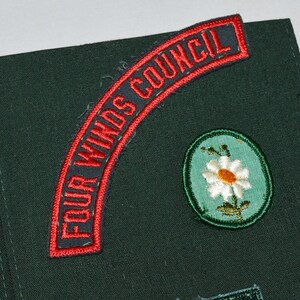 May include: A green fabric patch with a red and white embroidered patch that says "Four Winds Council". There is also a green and white embroidered patch of a daisy.