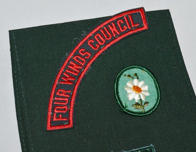 May include: A green fabric patch with a red and white embroidered patch that says "Four Winds Council". There is also a green and white embroidered patch of a daisy.