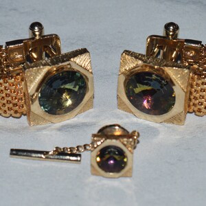 May include: A set of gold cufflinks with a chain and a tie tack. Each piece features a square-shaped setting with a round, iridescent gemstone.