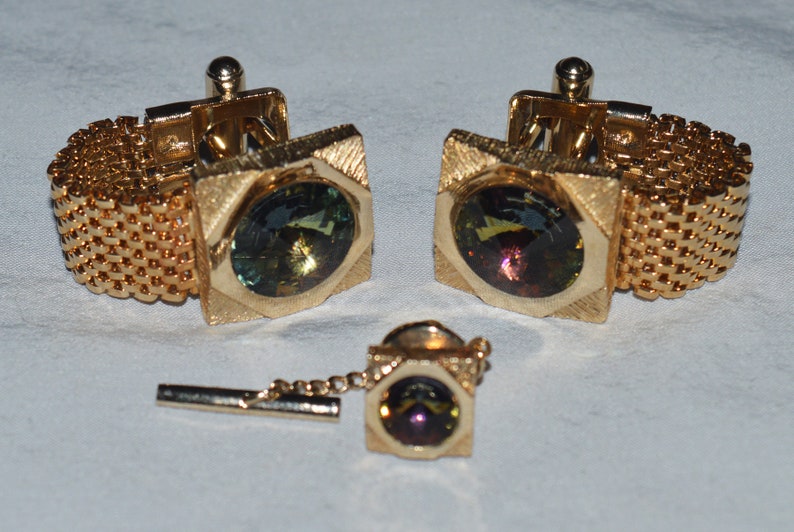 May include: A set of gold cufflinks with a chain and a tie tack. Each piece features a square-shaped setting with a round, iridescent gemstone.