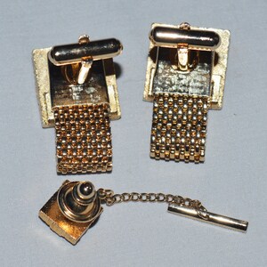 May include: A set of gold-tone cufflinks with a chain design and a matching tie tack. The cufflinks and tie tack have a square shape with a gold-tone finish.