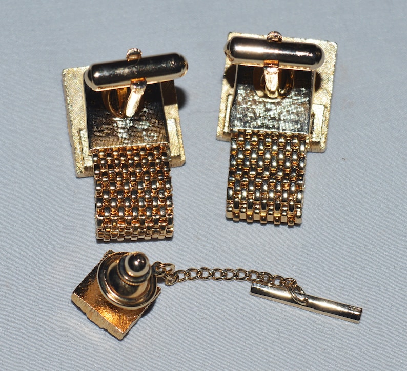 May include: A set of gold-tone cufflinks with a chain design and a matching tie tack. The cufflinks and tie tack have a square shape with a gold-tone finish.