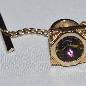 May include: A gold tie tack with a square frame and a round, iridescent purple and green gemstone in the center. The tie tack has a chain attached to a bar.