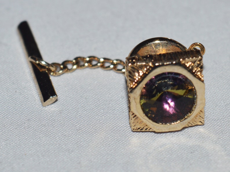 May include: A gold tie tack with a square frame and a round, iridescent purple and green gemstone in the center. The tie tack has a chain attached to a bar.